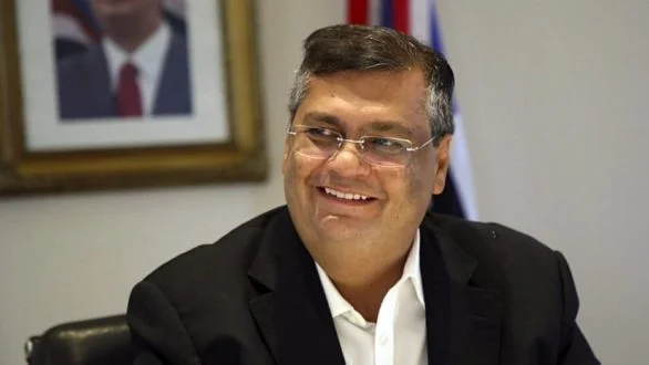 um homem de terno sentado em frente a uma bandeira.
